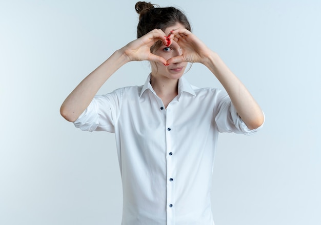 Young confident blonde russian girl gestures and looks through heart hand sign isolated on white space with copy space