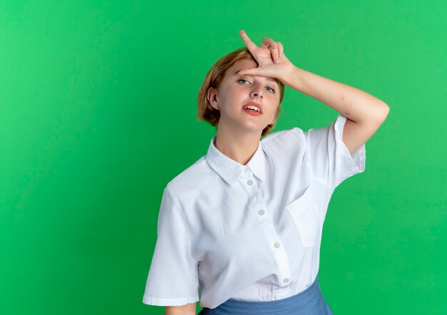 Young confident blonde russian girl gestures loser sign isolated on green background with copy space