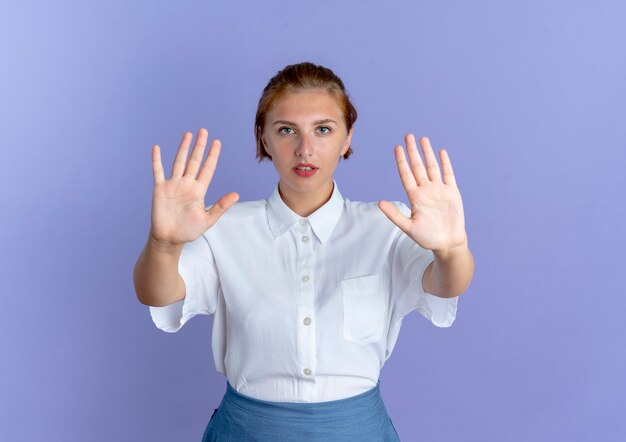 Young confident blonde russian girl  gestures stop with two hands isolated on purple background with copy space