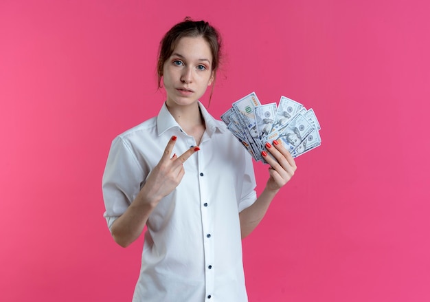 Young confident blonde russian girl gestures victory hand sign holding money on pink  with copy space