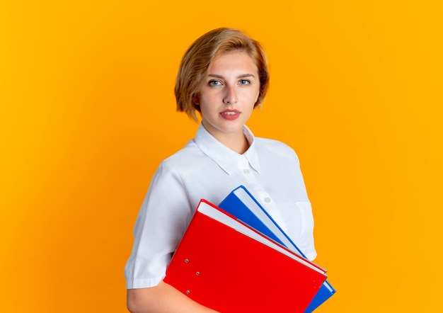 Young confident blonde russian girl holds file folders looking at camera isolated on orange background with copy space