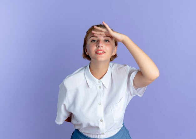 Young confident blonde russian girl holds hand to head trying to see isolated on purple background with copy space