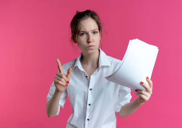 Free photo young confident blonde russian girl holds paper sheets pointing up isolated on pink space with copy space