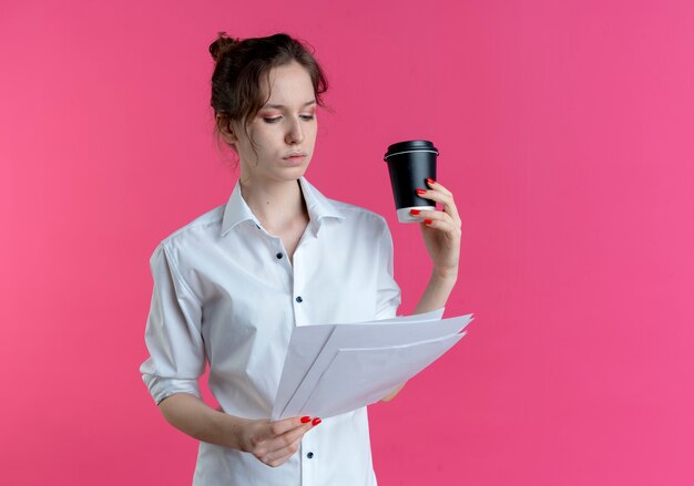 若い自信を持って金髪のロシアの女の子は、コピースペースとピンクのスペースで隔離のコーヒーカップを保持している紙シートを見て