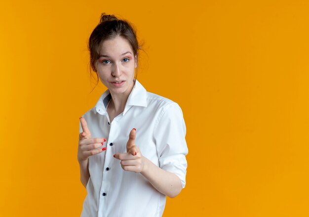 young confident blonde russian girl looks and points at camera isolated on orange background