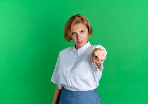 Young confident blonde russian girl points at camera isolated on green background with copy space