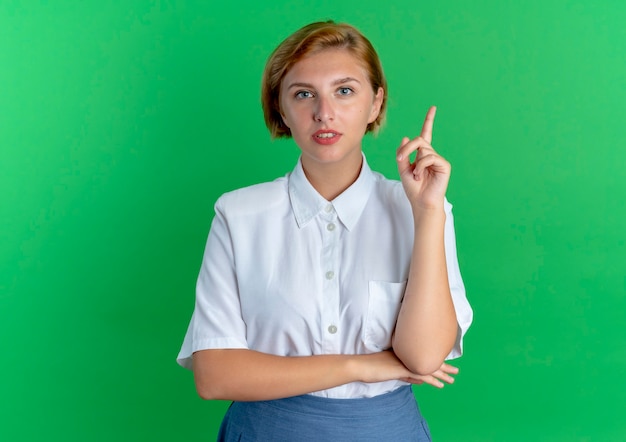 Young confident blonde russian girl points up looking at camera isolated on green background with copy space