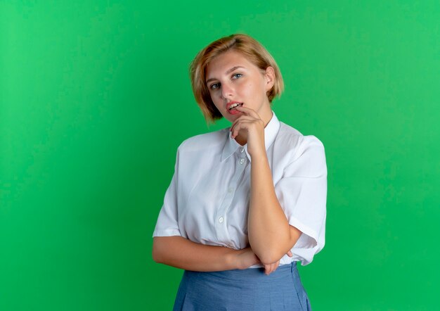 Young confident blonde russian girl puts finger on lips isolated on green background with copy space