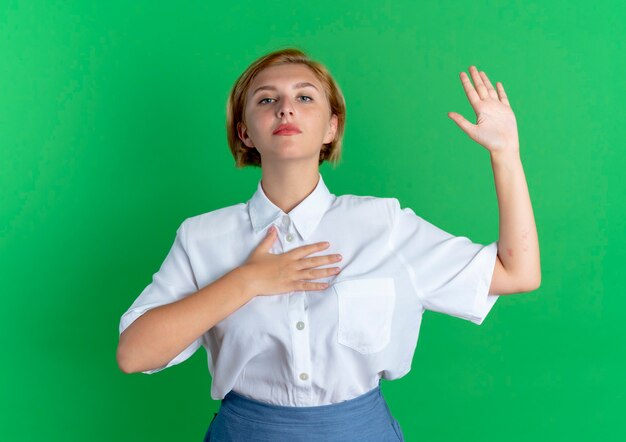 Молодая уверенная в себе русская блондинка кладет руку на грудь и поднимает руку, изолированную на зеленом фоне с копией пространства