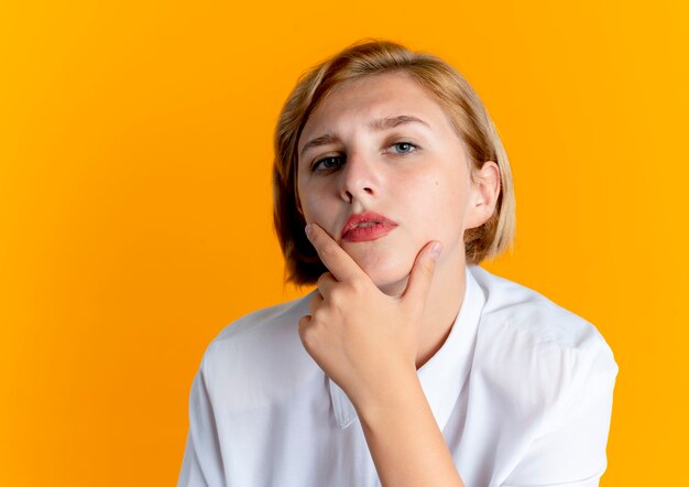 Young confident blonde russian girl puts hand on chin isolated on orange background with copy space