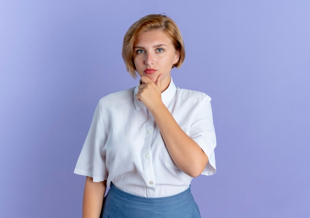 Young confident blonde russian girl puts hand on chin isolated on purple background with copy space