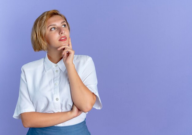 Young confident blonde russian girl puts hand on chin looking at side isolated on purple background with copy space