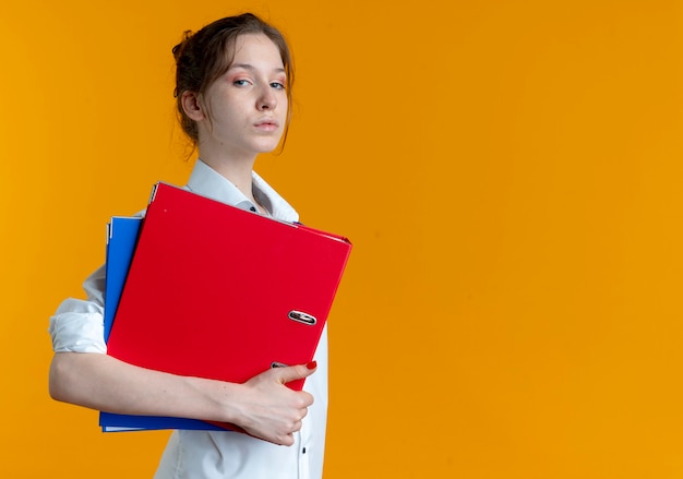 Young confident blonde russian girl stands sideways holding file folders on orange  with copy space