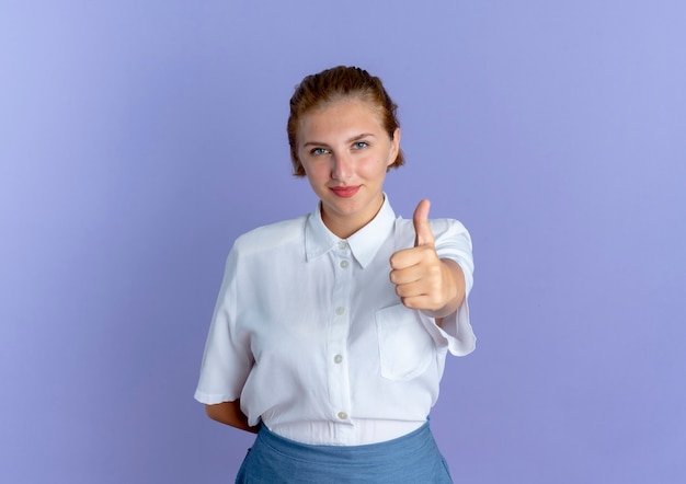 若い自信を持って金髪のロシアの女の子は、コピースペースで紫色の背景に分離されたカメラを見て親指を立てる
