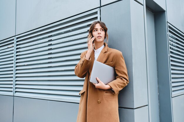 Young confident casual businesswoman in coat with laptop talking on cellphone outdoor