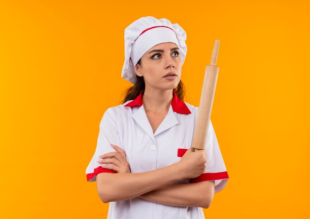 Молодая уверенная кавказская девушка-повар в униформе шеф-повара скрещивает руки и держит скалку на оранжевом фоне с копией пространства