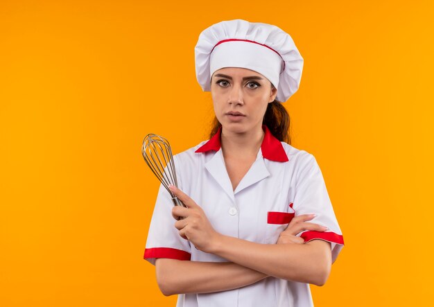 Young confident caucasian cook girl in chef uniform crosses arms and holds whisk isolated on orange background with copy space
