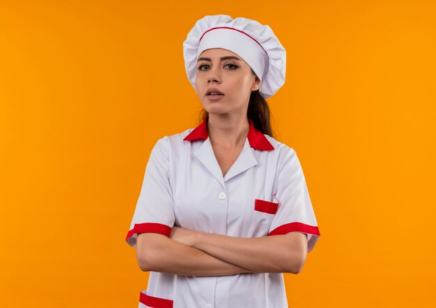 요리사 유니폼에 젊은 자신감 백인 요리사 소녀 팔을 교차하고 복사 공간이 오렌지 배경에 고립 된 카메라를 찾습니다
