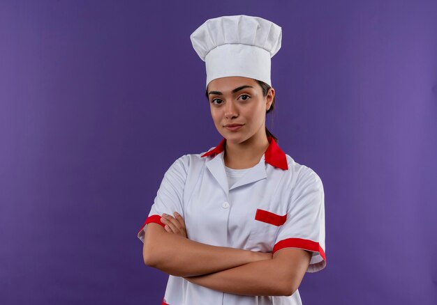 Young confident caucasian cook girl in chef uniform crosses arms and looks at camera isolated on violet wall with copy space
