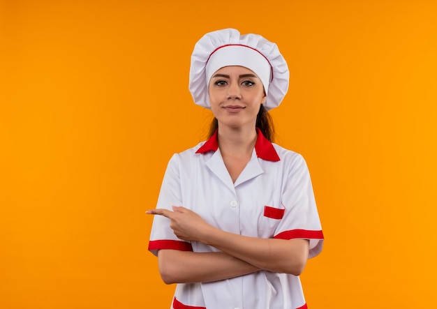 Young confident caucasian cook girl in chef uniform crosses arms and points to the side isolated on orange wall with copy space