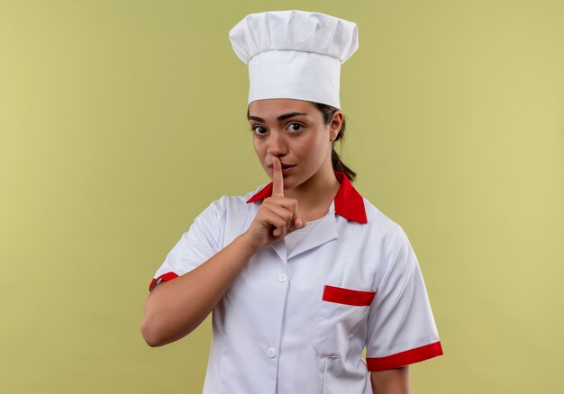Молодая уверенная кавказская девушка-повар в униформе шеф-повара жестами молчит знак рукой, изолированной на зеленой стене с копией пространства