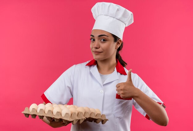 Молодая уверенная в себе кавказская девушка-повар в униформе шеф-повара держит партию яиц и показывает палец вверх, изолированную на розовой стене с копией пространства