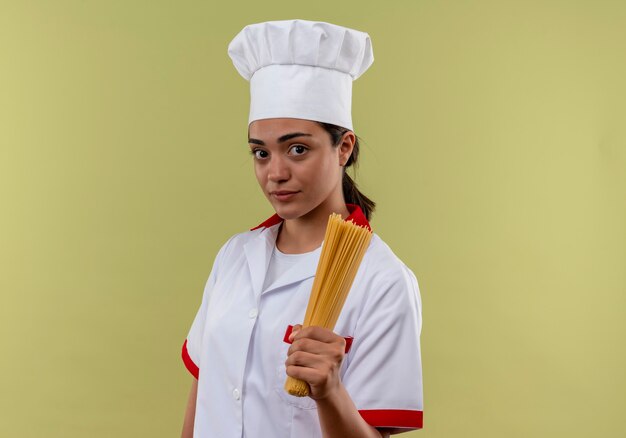 Young confident caucasian cook girl in chef uniform holds bunch of spaghetti isolated on green wall with copy space