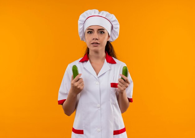 Young confident caucasian cook girl in chef uniform holds cucumbers in both hands isolated on orange wall with copy space