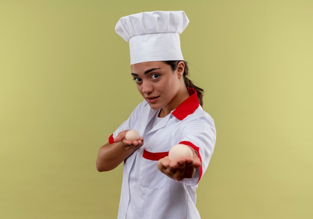 La giovane ragazza caucasica sicura del cuoco in uniforme del cuoco unico tiene fuori le uova isolate sulla parete verde con lo spazio della copia