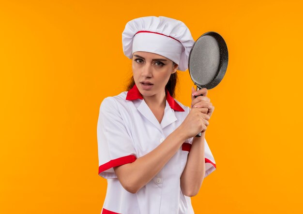 요리사 유니폼에 젊은 자신감 백인 요리사 소녀 복사 공간 오렌지 벽에 고립 된 양손으로 프라이팬을 보유
