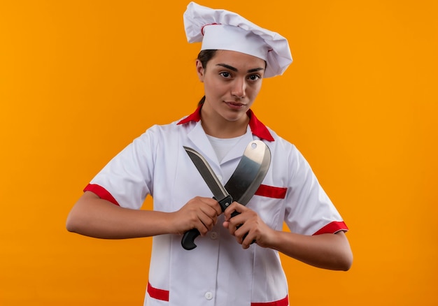 La giovane ragazza caucasica sicura del cuoco in uniforme del cuoco unico tiene i coltelli isolati sulla parete arancione con lo spazio della copia