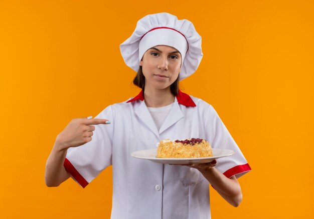 シェフの制服を着た若い自信を持って白人料理人の女の子は、コピースペースでオレンジ色の背景に分離されたプレート上のケーキを保持し、指さします