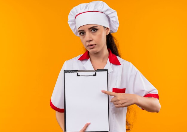 Young confident caucasian cook girl in chef uniform holds and points at clipboard isolated on orange wall with copy space