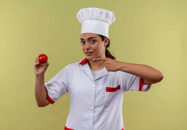 Young confident caucasian cook girl in chef uniform holds and points at tomato isolated on green wall with copy space