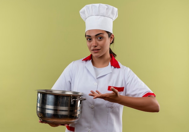 Young confident caucasian cook girl in chef uniform holds pot and points with hand isolated on green wall with copy space