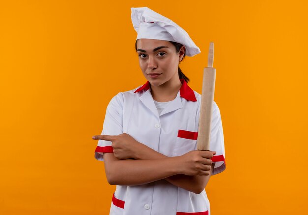 Young confident caucasian cook girl in chef uniform holds rolling pin and points to the side isolated on orange wall with copy space