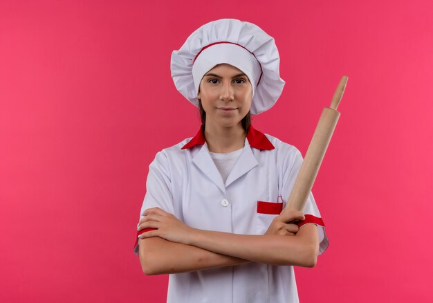 シェフの制服を着た若い自信を持って白人料理人の女の子は、コピースペースとピンクのスペースに分離された腕を組んで麺棒を保持します