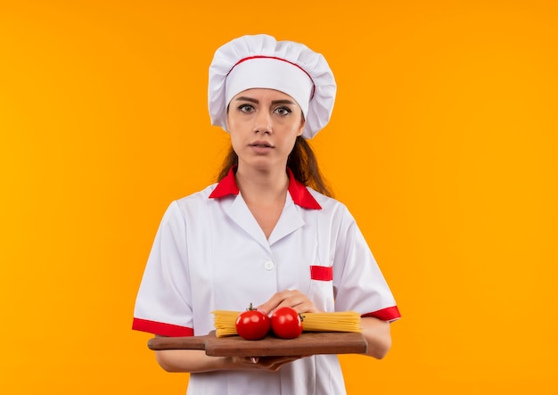 Young confident caucasian cook girl in chef uniform holds tomatoes and bunch of spaghetti on cutting board isolated on orange wall with copy space