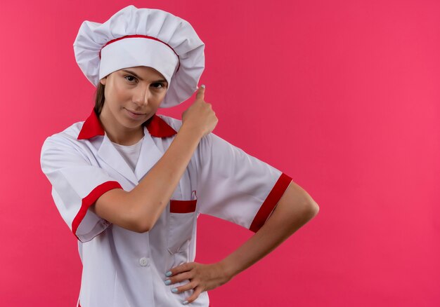 Young confident caucasian cook girl in chef uniform points back isolated on pink space with copy space