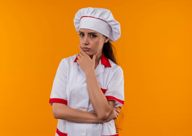 La giovane ragazza caucasica sicura del cuoco in uniforme del cuoco unico mette la mano sul mento isolata sulla parete arancione con lo spazio della copia