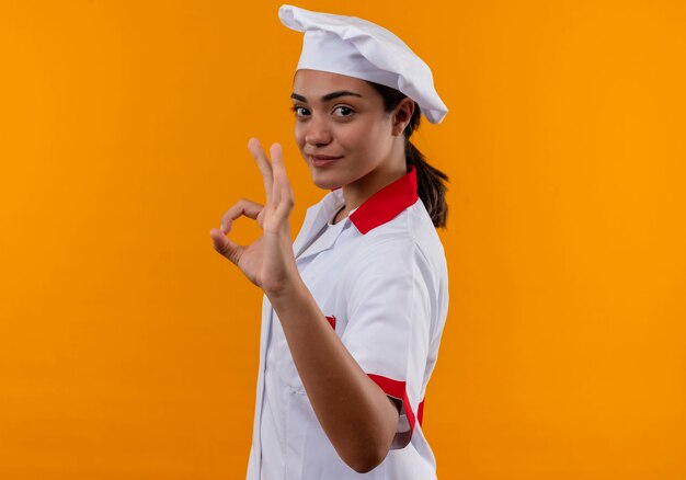 Молодая уверенная в себе кавказская девушка-повар в униформе шеф-повара стоит боком и жестикулирует знак рукой на оранжевой стене с копией пространства