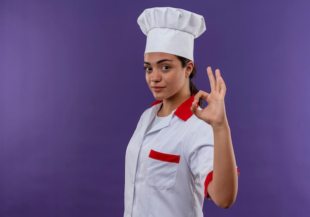 Young confident caucasian cook girl in chef uniform stands sideways and gestures ok hand sign isolated on violet wall with copy space