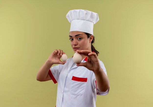 Young confident caucasian cook girl in chef uniform stands sideways and holds eggs isolated on green wall with copy space