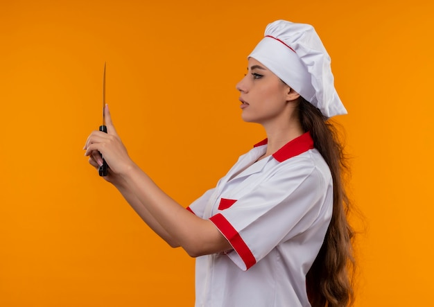 シェフの制服を着た若い自信を持って白人料理人の女の子は横に立って、コピースペースでオレンジ色の壁に分離されたナイフを保持します