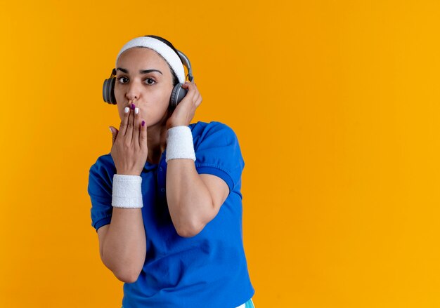 Young confident caucasian sporty woman wearing headband and wristbands on headphones sends kiss with hand on orange  with copy space