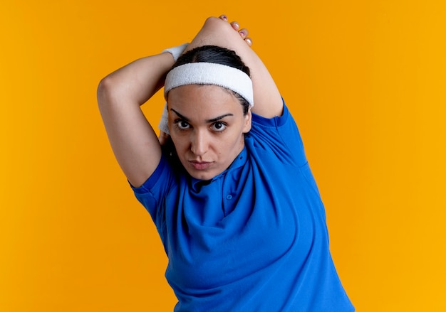 Молодая уверенная кавказская спортивная женщина с повязкой на голову и браслетами держит руку за спиной, изолированную на оранжевом фоне с копией пространства