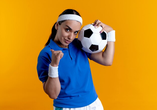 молодая уверенная в себе кавказская спортивная женщина с повязкой на голову и браслетами держит мяч, указывая назад