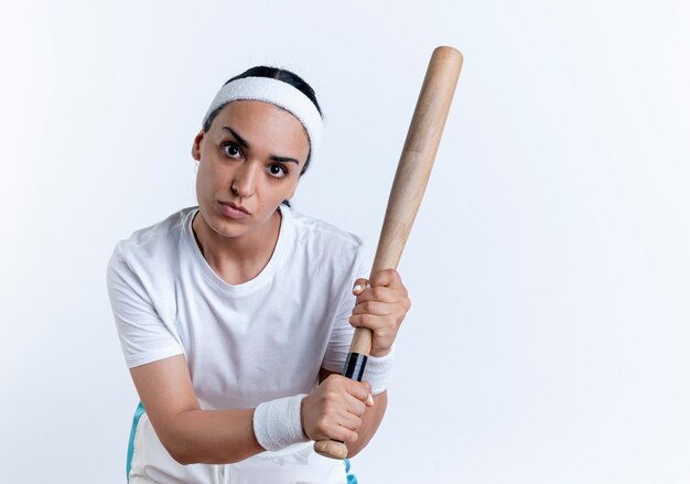 young confident caucasian sporty woman wearing headband and wristbands holds bat looking at camera