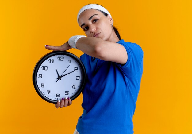 Молодая уверенная кавказская спортивная женщина с повязкой на голову и браслетами держит часы, изолированные на оранжевом фоне с копией пространства
