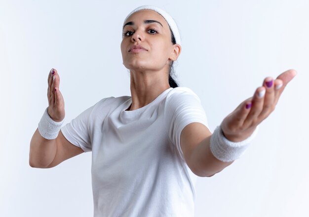 Young confident caucasian sporty woman wearing headband and wristbands holds hands out isolated on white space with copy space